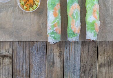 GỎI CUỐN và GỎI ĐU ĐỦ Auf einem Holztisch liegen auf einem Backpapier drei frisch gerollte Sommerrollen. Daneben steht eine Soße zum Dippen.