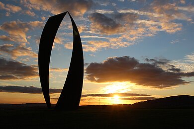 Kunstwerk in Form eines geschwungenen O vor blauem Himmel mit Sonnenuntergangsstimmung im Hintergrund