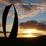 Kunstwerk in Form eines geschwungenen O vor blauem Himmel mit Sonnenuntergangsstimmung im Hintergrund