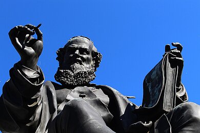 Das Bild zeigt die Bronzestatue eines Mannes, der gerade die Hand hebt. Die Statue stellt Hans Sachs dar. Im Hintergrund blauer Himmel