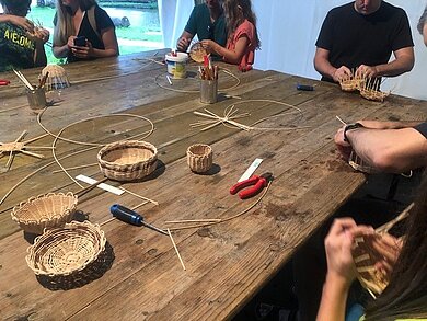 An einem großen Holztisch sitzen Menschen, die mit dem Flechten eines Korbes beschäftigt sind. Auf dem Tisch befinden sich Materialien und Werkzeug zum Flechten von Körben.