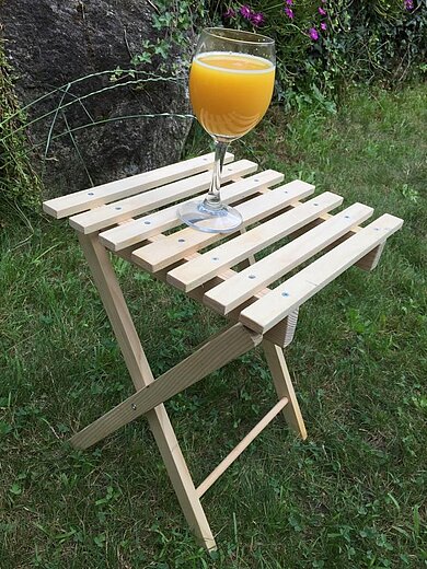 Zusammenklappbares Beistelltischchen aus Holz auf dem ein Glas mit orangener Flüssigkeit steht. 