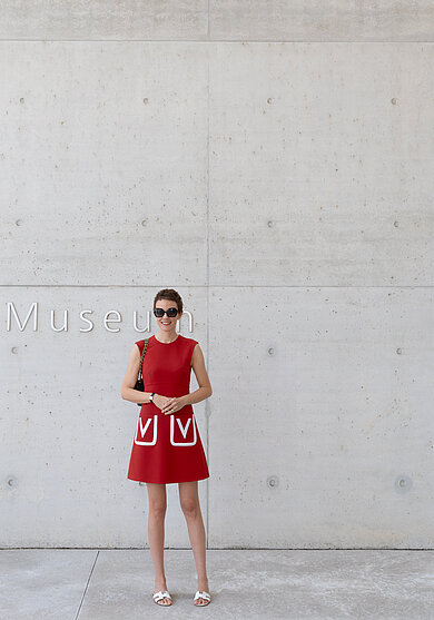 Frau in rotem ärmellosem Kleid mit weißen Taschen und weißen Sandalen steht vor einer hellgrauen Betonwand mit dem Schriftzug 'Museum'