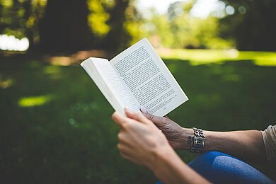 Person hält ein aufgeschlagenes Buch und liest im Freien bei Tageslicht

