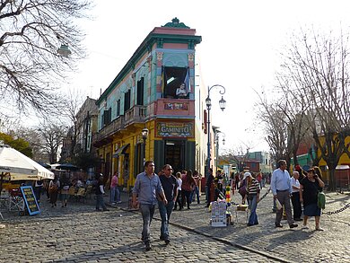 Straßenszene in Argentinien. Menschen spazieren über Kopfsteinpflaster, im Hintergrund steht ein buntes Haus.