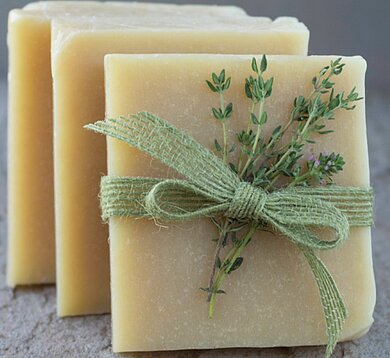 Drei hintereinander aufgestellte quadratische Seifen, im Vordergrund mit Kräutern verziert. 