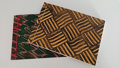 Zwei übereinander liegende Bücher mit unterschiedlichen Einbänden und Fadenheftung. Ein Bucheinband hat Wellen, der andere wirkt wie geflochten. 