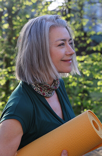 Eine Frau hält eine Yogamatte unter den Armen und lächelt in die Ferne.