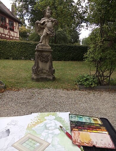Blick auf eine Statue im Garten, im Vordergrund eine gemalte Abbildung der Statue in Aquarellfarben, ein Aquarellmalkasten und Pinsel.