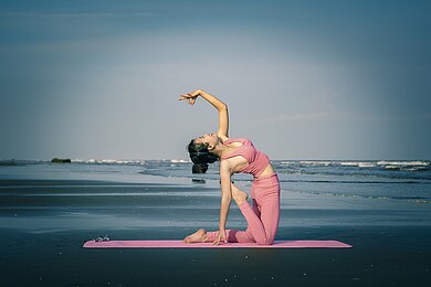 Eine Frau führt auf einem Strand eine Yogapose aus. Hinter ihr ist das Meer zu sehen.