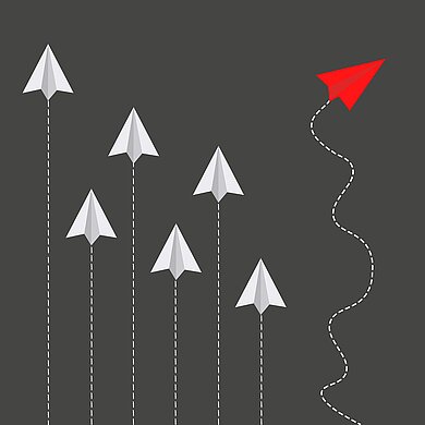 Kreativmethoden für innovatives Arbeiten  Mehrere Papierflieger fliegen gerade aus. Ein roter Papierflieger fliegt aus der Reihe.